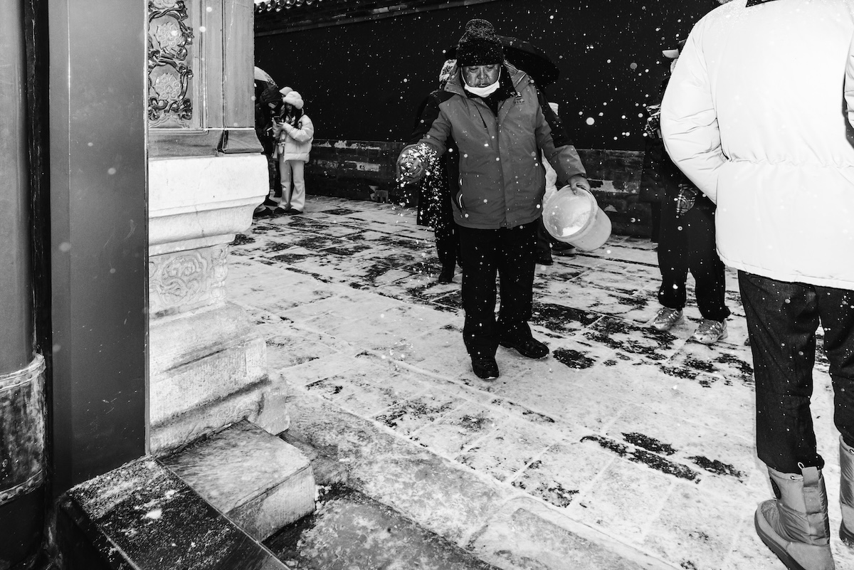 An elderly woman in a dark coat and white mask brushes snow from a stone step.